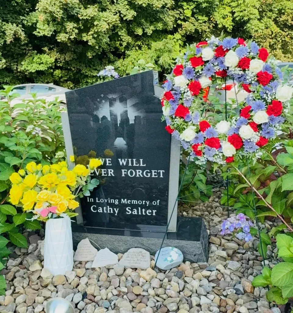 A red, white and blue wreath beside Cathy's memorial stone, which reads "We Will Never Forget, In Loving Memory of Cathy Salter", with yellow roses in a white vase.