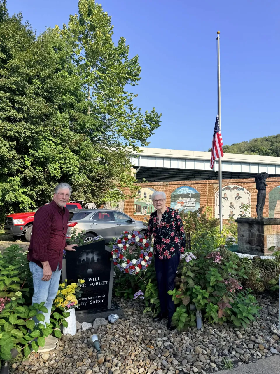 Two family members stand on either side of Cathy's memorial stone and the new wreath, with the American flag at half-staff and the Lisbon Street murals behind them.
